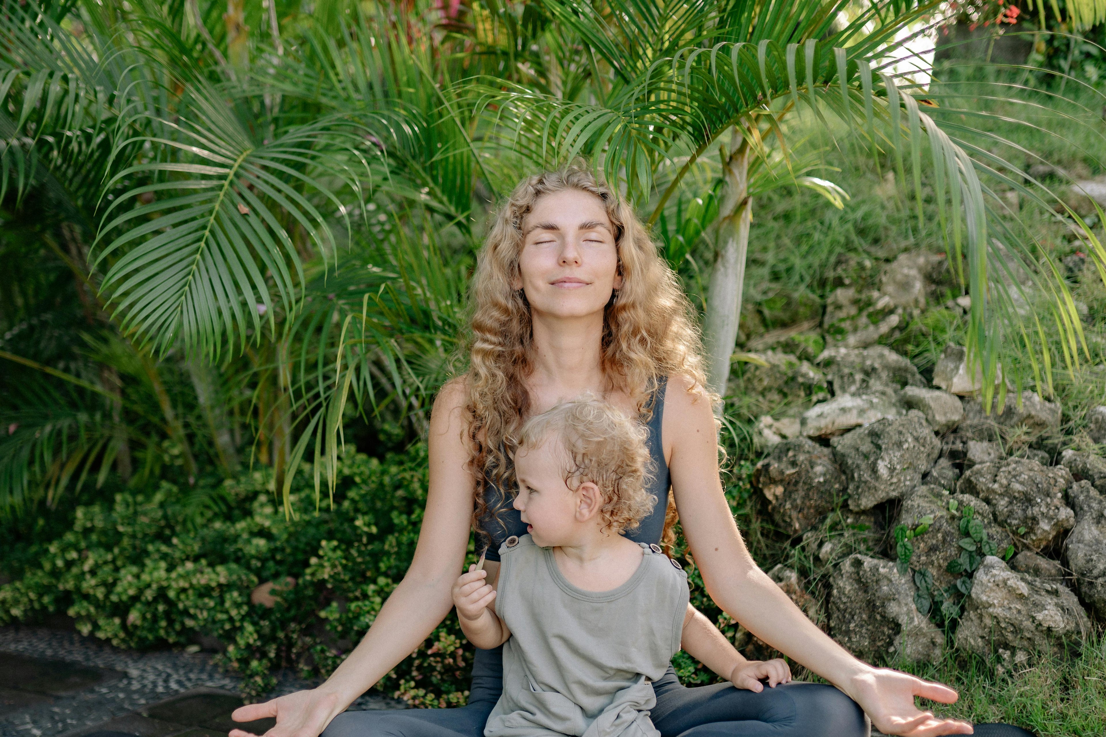 Woman and child sitting on a yoga mat outdoors with greenery in the background